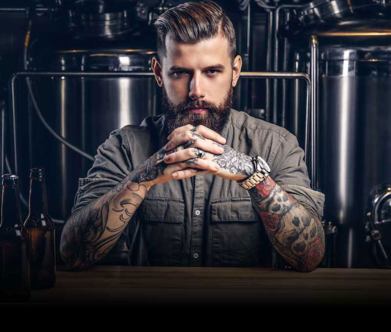 A bearded man with tattoos on his arms, sitting with his hands crossed and elbows on a bar, with beer bottles nearby and large silver stills in the background.