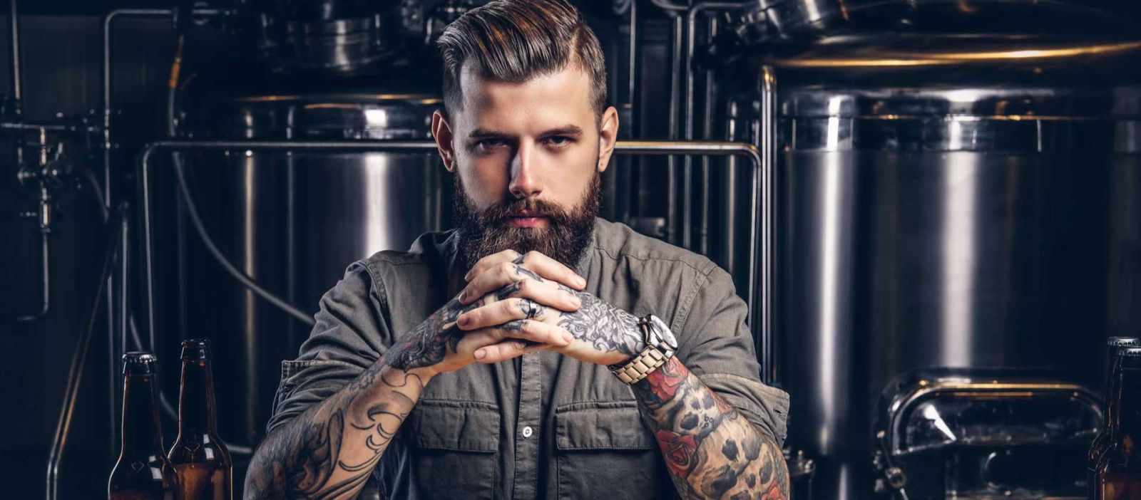 A bearded man with tattoos on his arms, sitting with his hands crossed and elbows on a bar, with beer bottles nearby and large silver stills in the background.