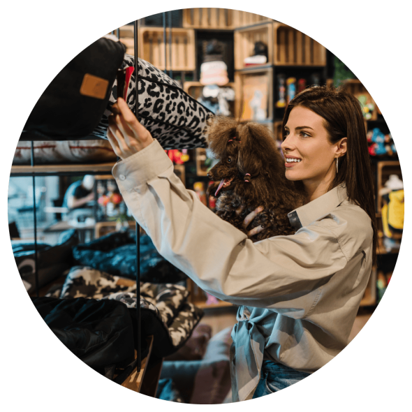 A woman holds her pet dog as she browses a pet store and checks out a tag on a leopard skin patterned pillow.