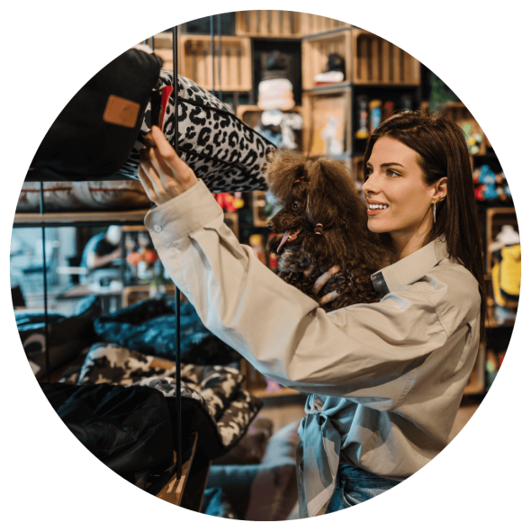 A woman holds her pet dog as she browses a pet store and checks out a tag on a leopard skin patterned pillow.